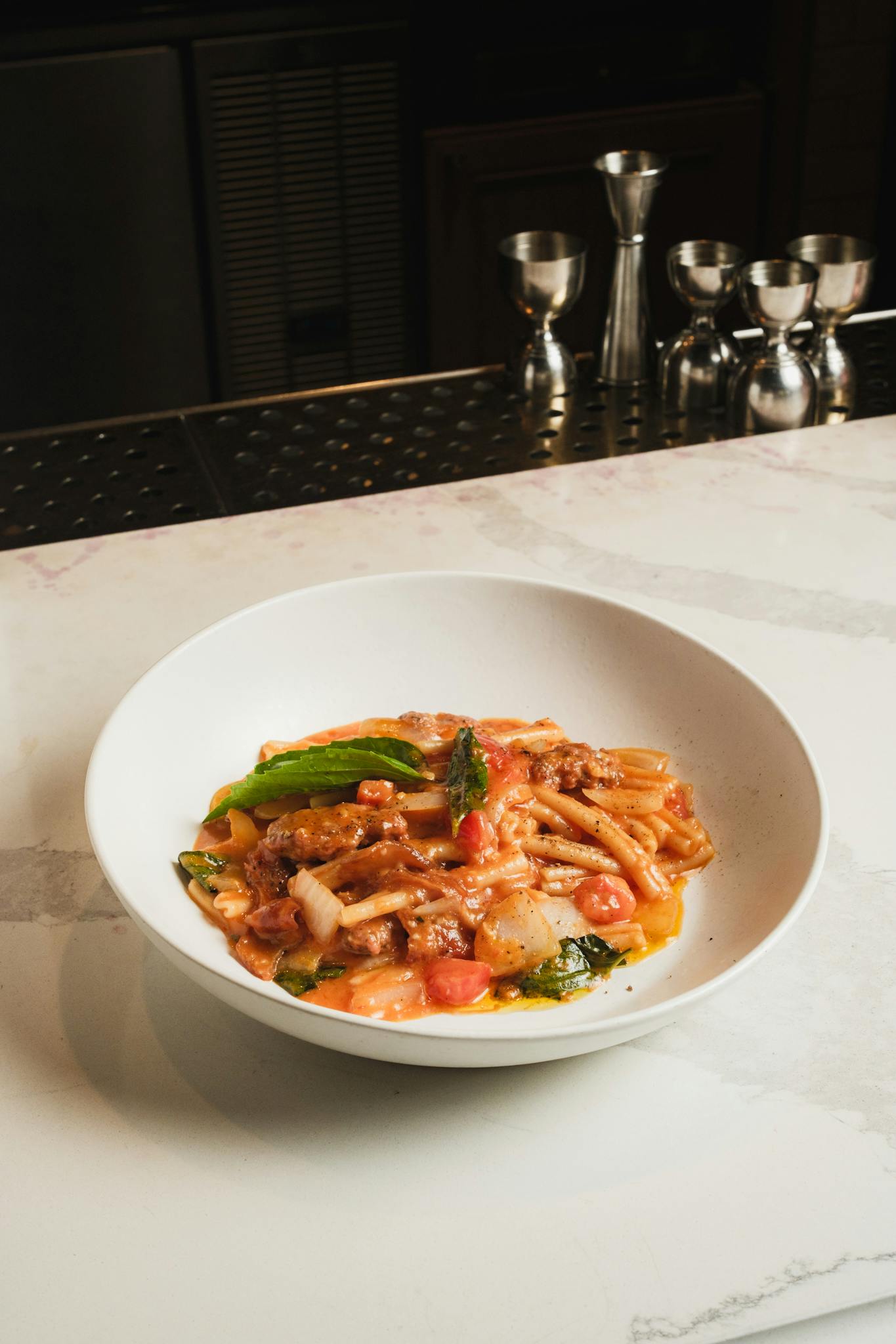 A bowl of savory pasta with meat, tomatoes, and greens served on a marble countertop.